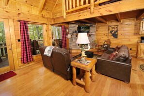 A cozy cabin interior with a wooden floor, two leather sofas, a small wooden table with a lamp, and a stone fireplace.