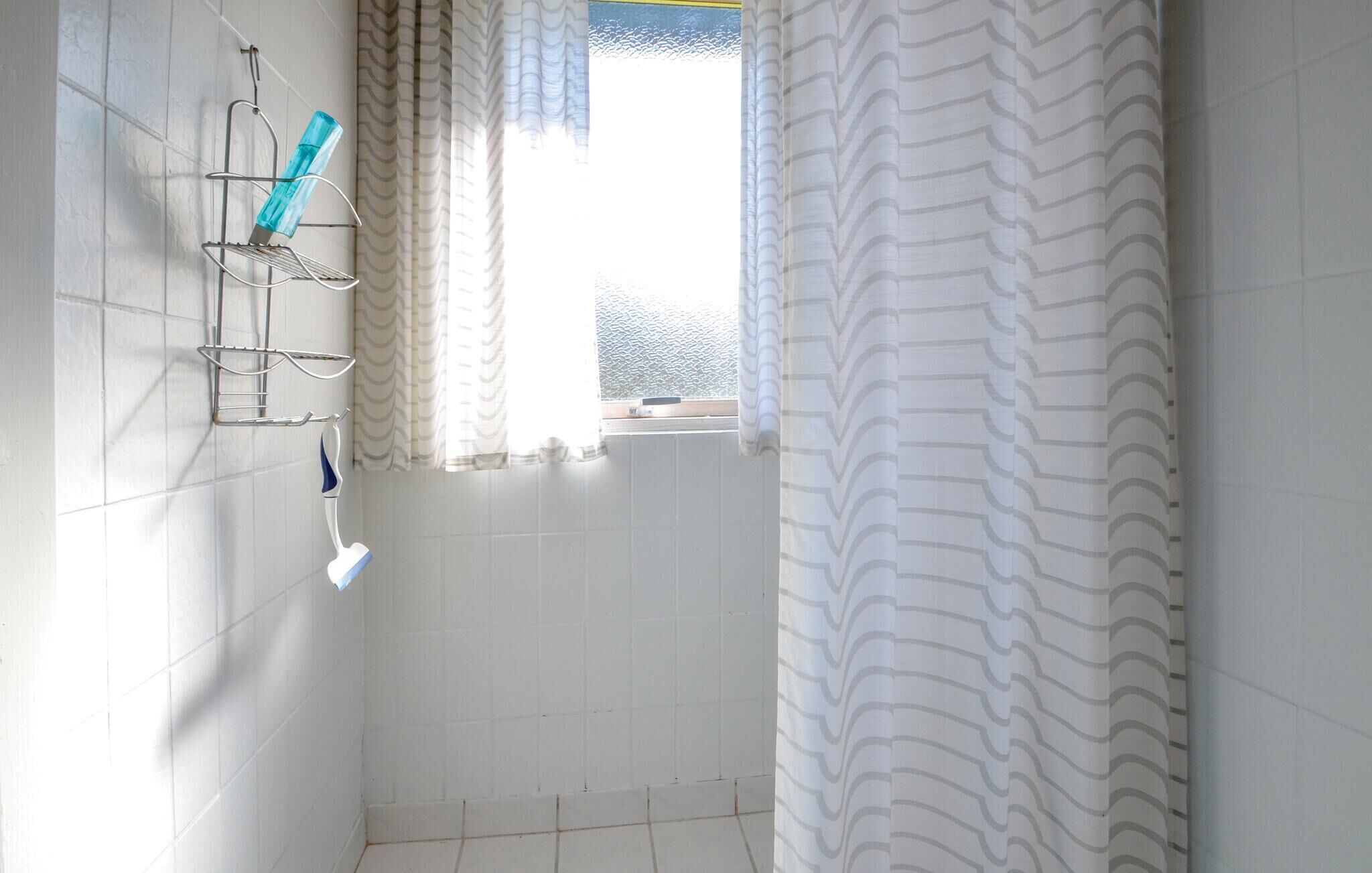 Un baño pequeño con paredes de azulejos blancos, cortina de ducha y una ventana con cortinas.