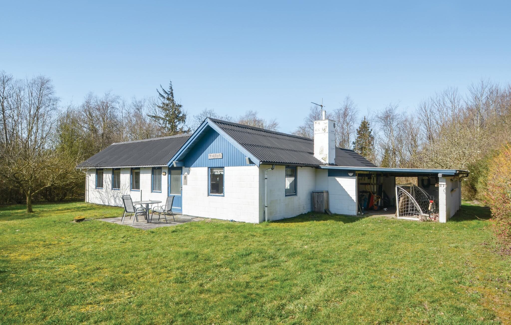 Ein einstöckiges Haus mit blauem Dach und weißen Wänden, mit einer Terrasse mit Stühlen und Tisch, umgeben von einem grünen Rasen und Bäumen.