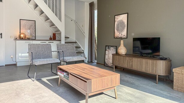 A modern living room with a staircase, grey upholstered chairs, a wooden coffee table, a TV, and artwork on the walls. - 3 bedroom stunning home in Hoeven (Hoeven)
