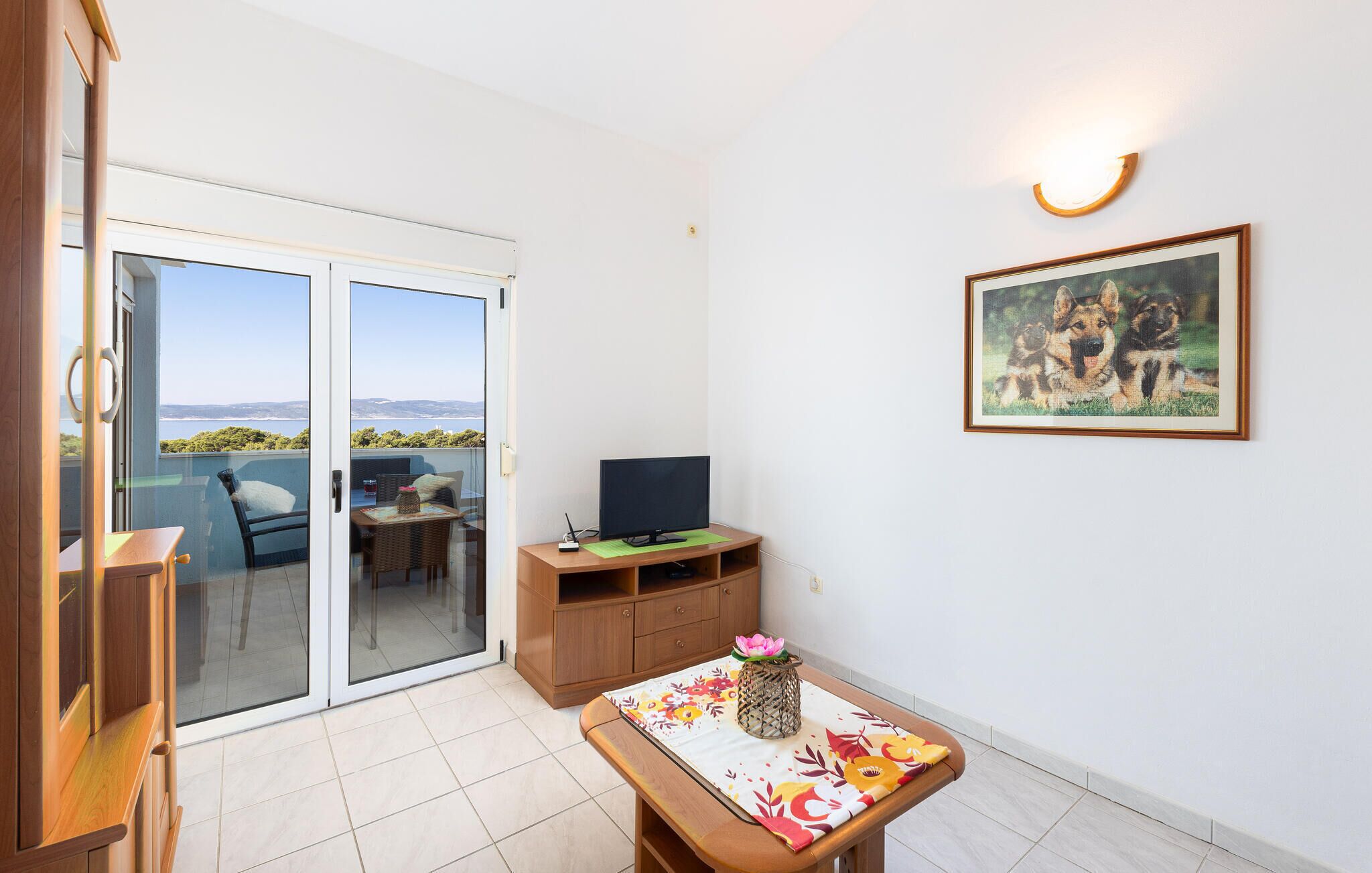 Una habitación con una puerta corrediza de vidrio que da a un balcón, un televisor sobre un mueble de madera, un cuadro enmarcado en la pared y una mesita con un jarrón.