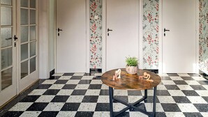 Um quarto com piso xadrez, três portas com papel de parede floral, uma mesa redonda de madeira com uma planta e figuras de madeira, e uma porta de vidro com painéis jateados.