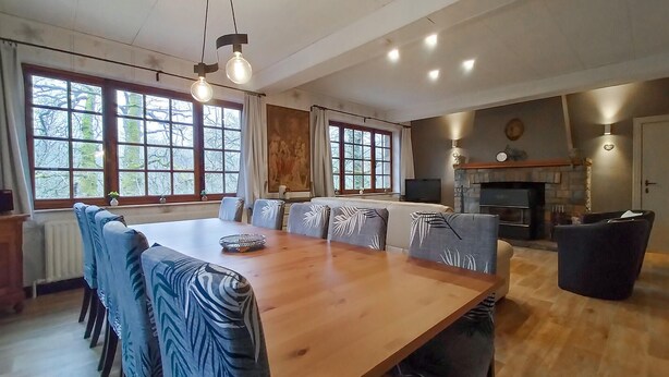 Uma sala de jantar espaçosa com mesa de madeira e cadeiras azuis estampadas, grandes janelas com cortinas e uma lareira ao fundo.