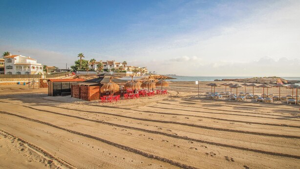 Une plage bordée de rangées de transats et de parasols, un petit bâtiment avec un toit de chaume, et une rangée de maisons en arrière-plan.