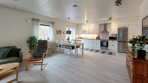 A spacious living area with a dining table, chairs, a sofa, and a rocking chair. A kitchen with white cabinets and appliances is visible in the background.
