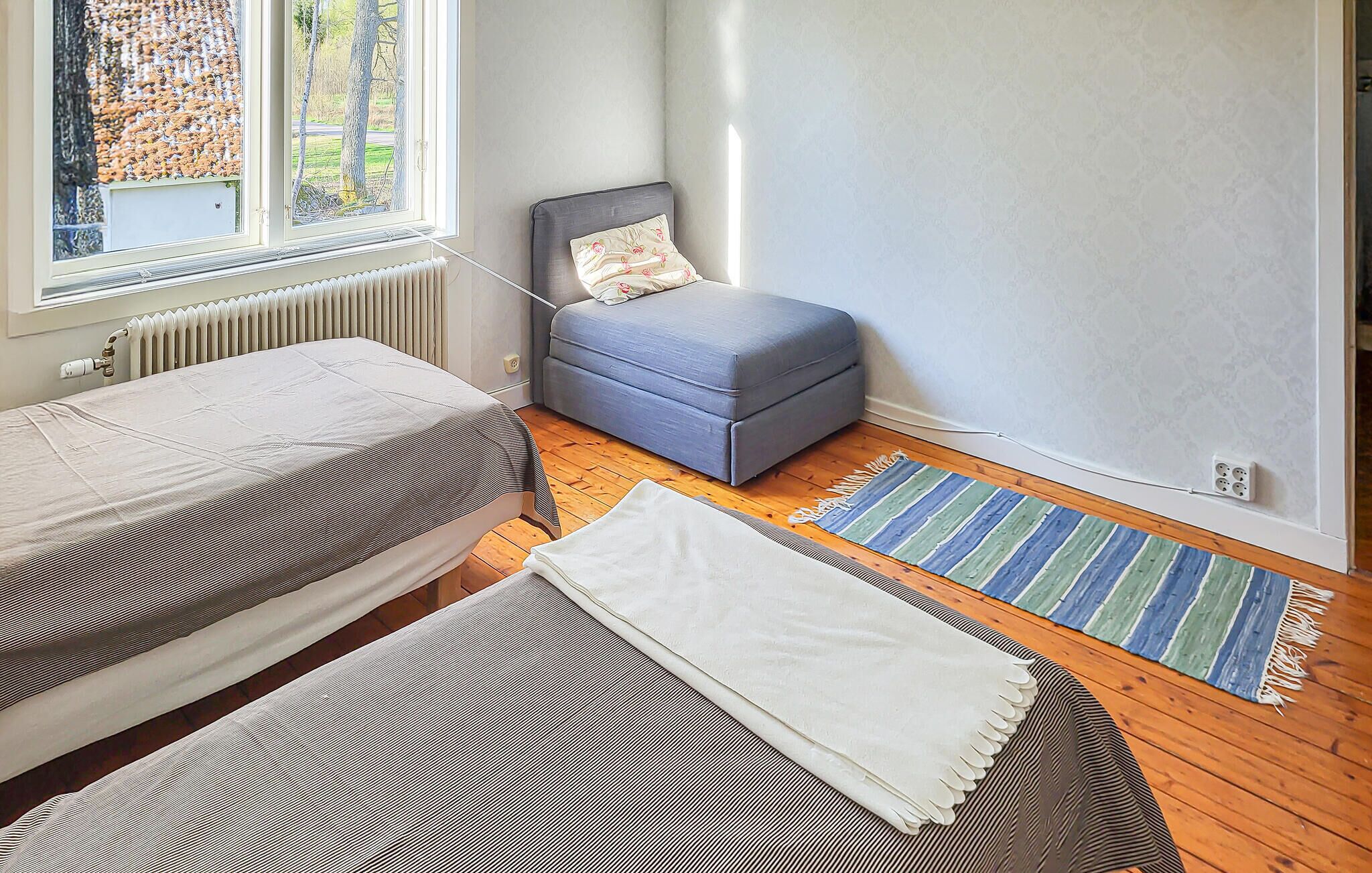 A bedroom with a bed, a grey sofa, a window with a view of trees, and a radiator.