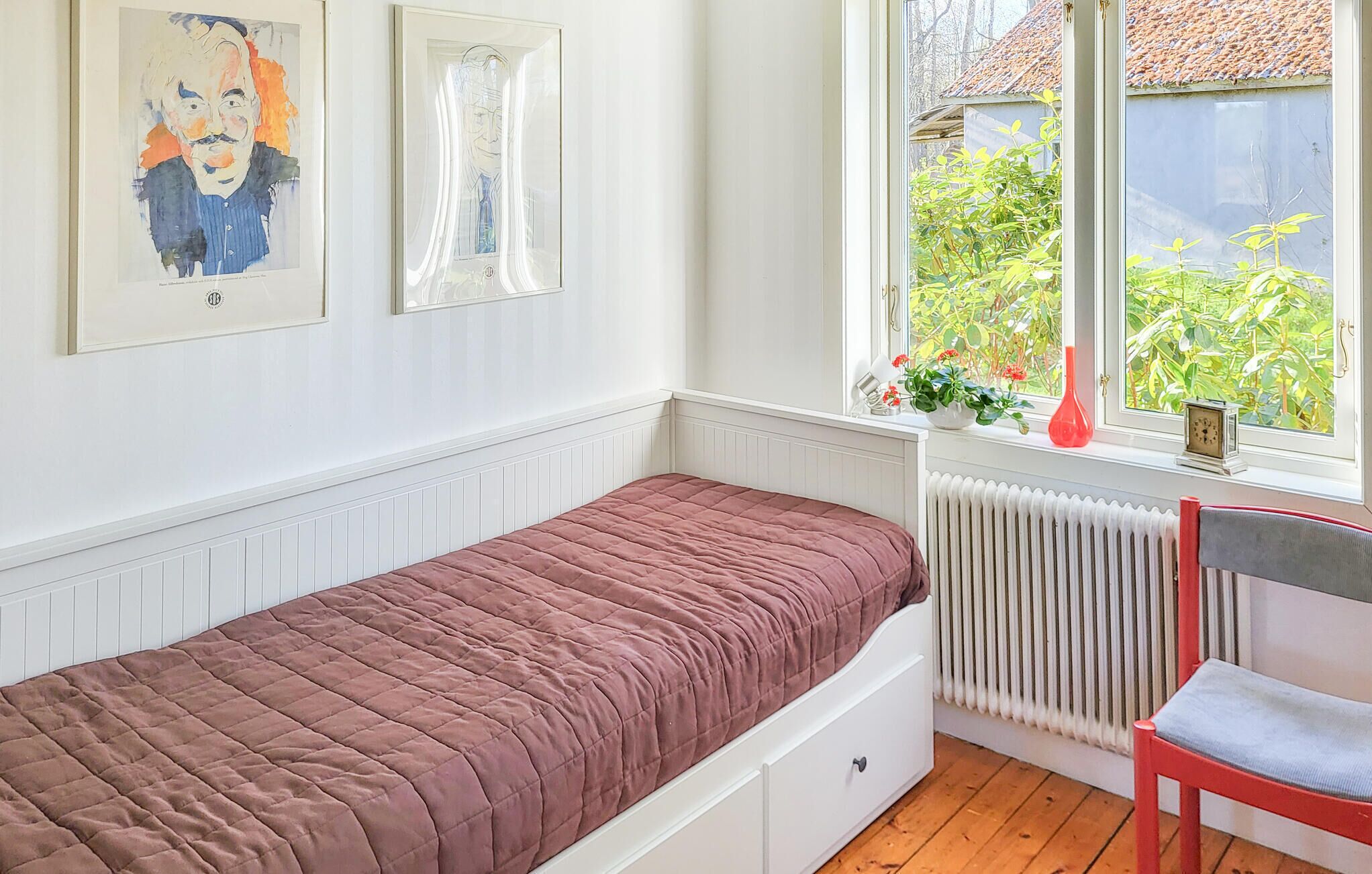 Une petite chambre avec un lit simple, une fenêtre, un radiateur et deux œuvres d’art encadrées accrochées au mur.