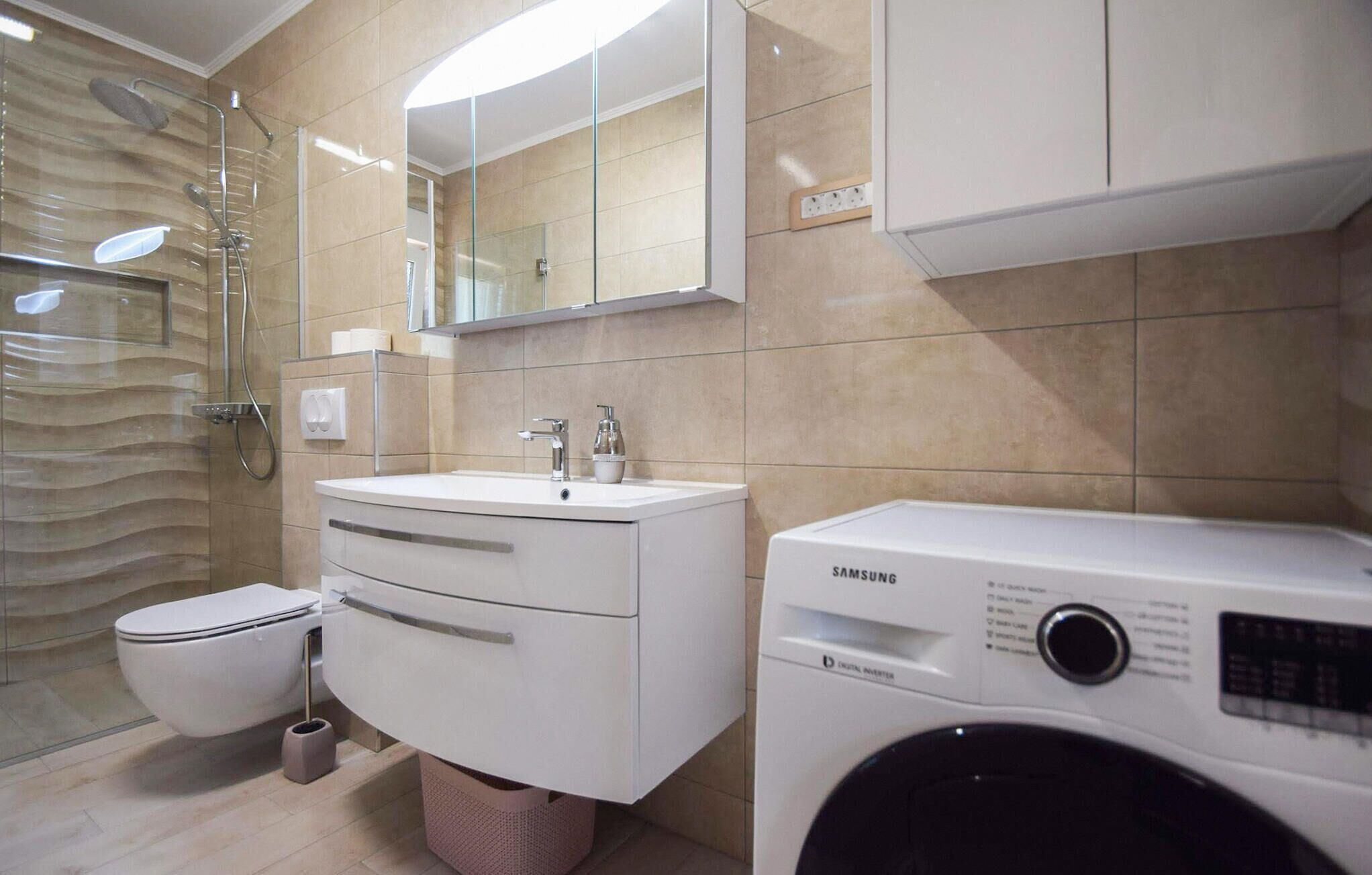 Un baño moderno con cabina de ducha de vidrio, un lavamanos blanco con espejo y una lavadora Samsung.