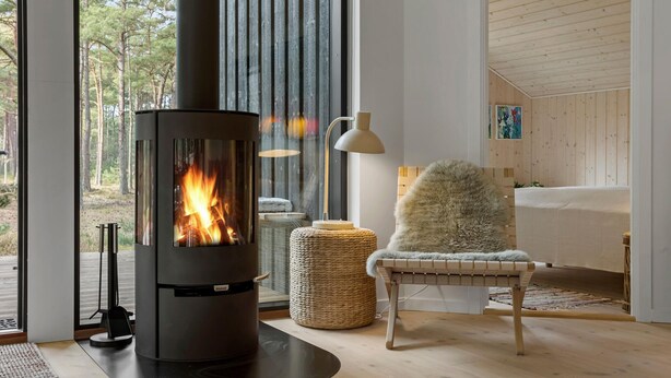 Ein modernes Wohnzimmer mit Kaminofen, großem Fenster, gewebtem Hocker und einem kuscheligen Sessel.