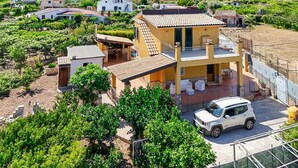 A two-story house with a balcony, surrounded by greenery and a parked SUV.