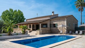 A two-story stone house with a swimming pool, outdoor seating, and a large tree.