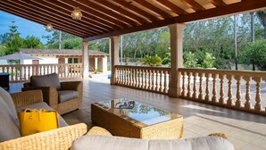 A covered outdoor seating area with wicker furniture and a glass coffee table, overlooking a garden and a building with a tiled roof.