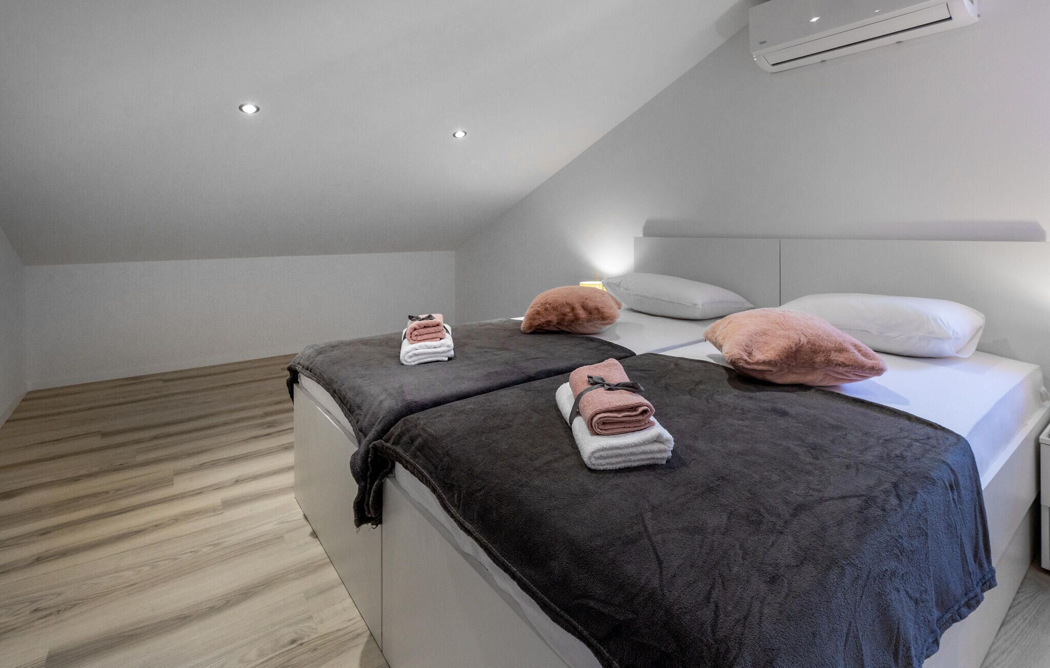 Un dormitorio moderno con techo inclinado, dos camas con ropa de cama gris oscuro y una mesita de noche pequeña.