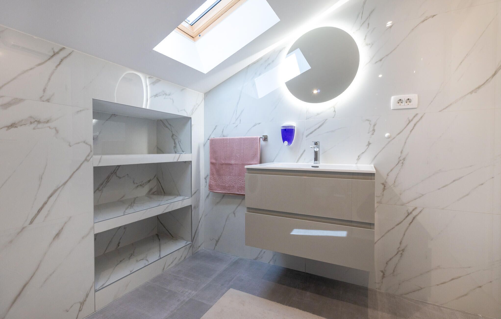 A modern bathroom with marble walls, a skylight, a vanity with a mirror, and a pink towel.
