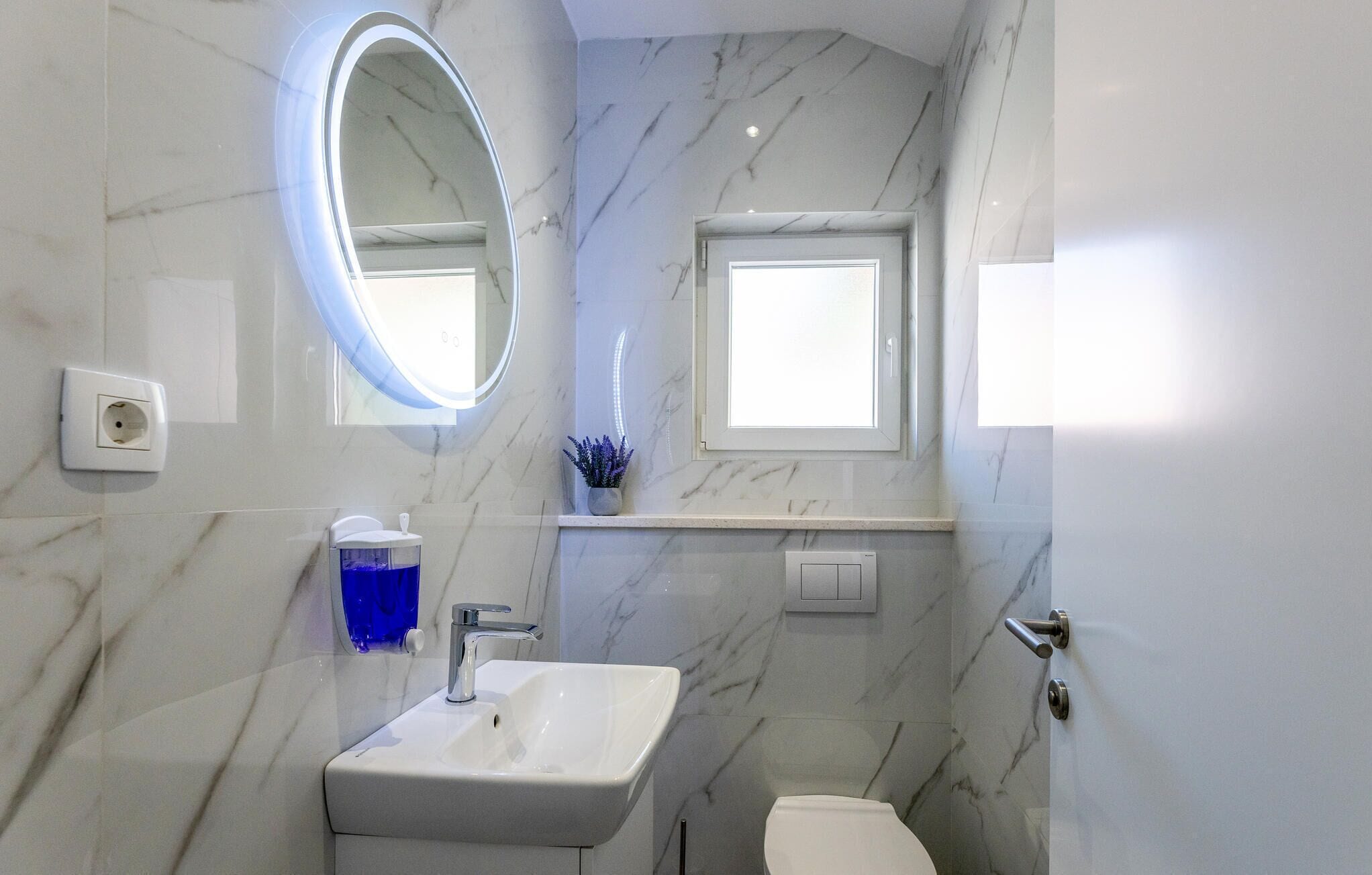 A bathroom with a marble wall, a white sink, a mirror, a window, and a toilet.