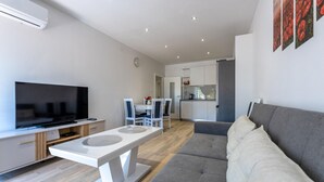 Una sala de estar moderna con un televisor de pantalla plana, un sofá gris y una zona de comedor con una mesa y sillas blancas.
