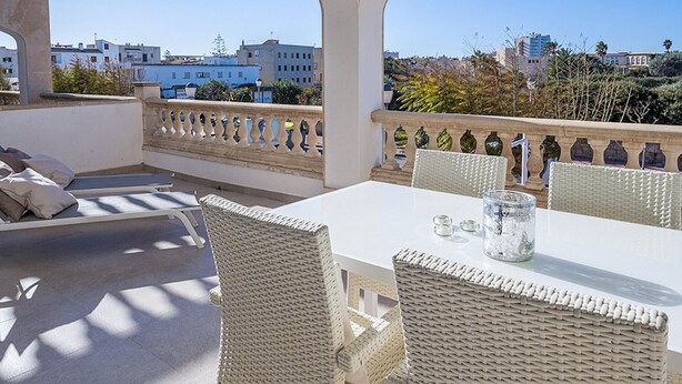 Una terraza con mobiliario de mimbre, una mesa con un florero de vidrio y vistas a edificios y árboles.