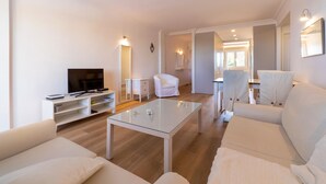 Una sala de estar moderna con un televisor de pantalla plana, sofás blancos, una mesa de centro de vidrio y un sillón blanco.