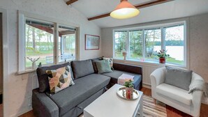 Ein gemütliches Wohnzimmer mit einer grauen Couch, einem weißen Sessel und einem Couchtisch. Große Fenster bieten einen Blick ins Freie.