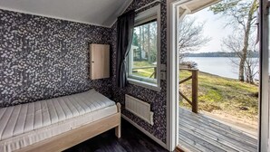 Ein kleines Zimmer mit einem Bett, einem Holzkasten und einem Fenster mit Blick auf einen See und Bäume.