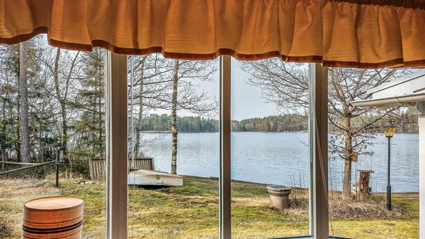 Ein Zimmer mit großem Fenster mit Blick auf einen See, einem Holzfass und einer Glasvase auf dem Fensterbrett.