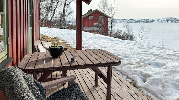 Un terrazzo in legno con tavolo e sedie, una pianta in vaso e vista su una casa rossa attraverso un paesaggio innevato.