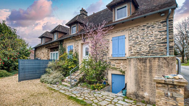 A stone house with a blue door and shutters, a small garden with flowering plants, and a stone pathway leading to the entrance.