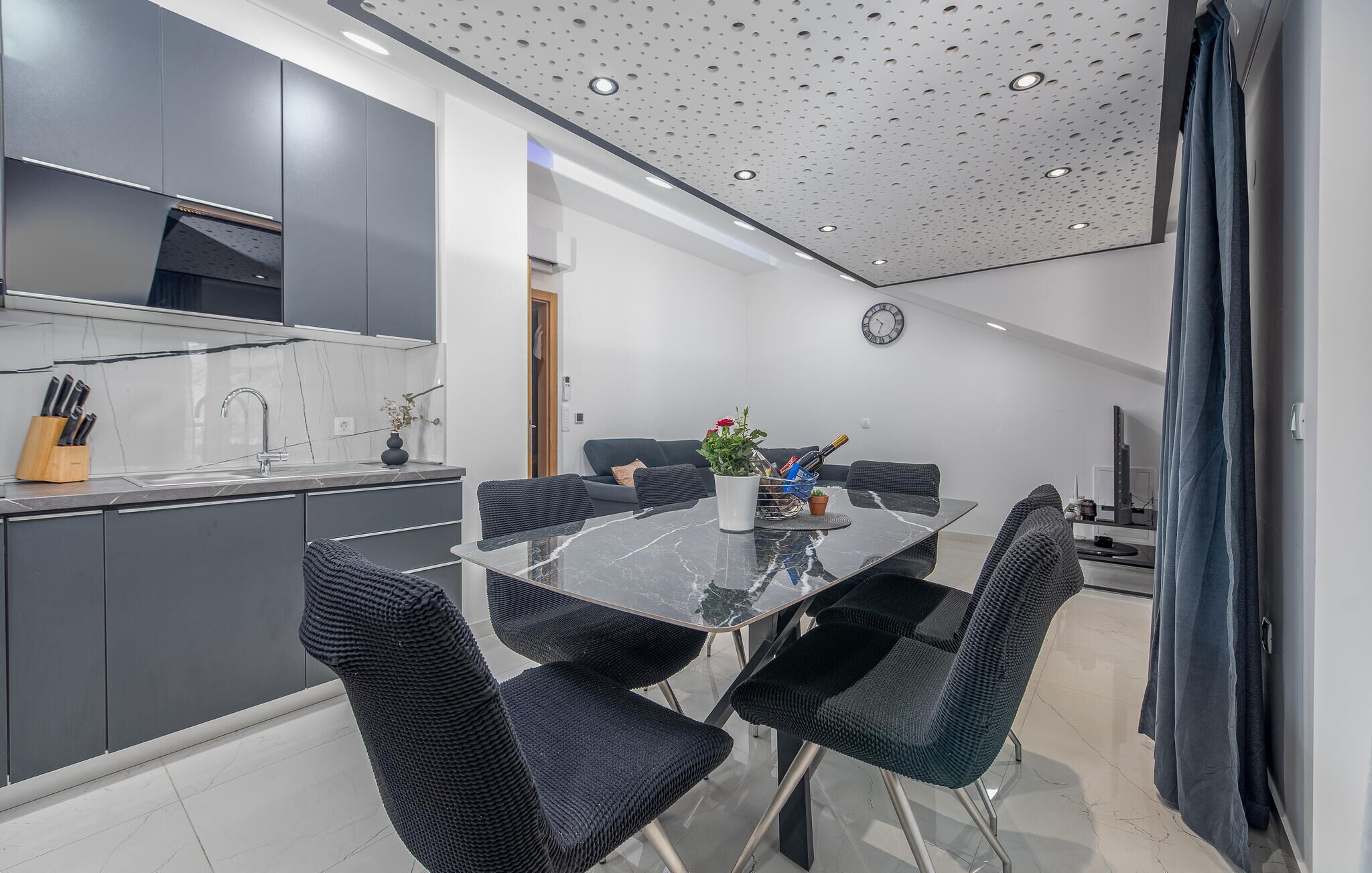 A modern kitchen with dark cabinets, a marble countertop, and a sink. There is a dining area with a glass table and black chairs.