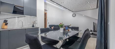 A modern kitchen with dark cabinets, a marble countertop, and a sink. There is a dining area with a glass table and black chairs.