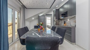 Un espace repas moderne avec une table en verre, des chaises en osier de couleur sombre et une cuisine dotée de meubles gris et d’appareils électroménagers en acier inoxydable.