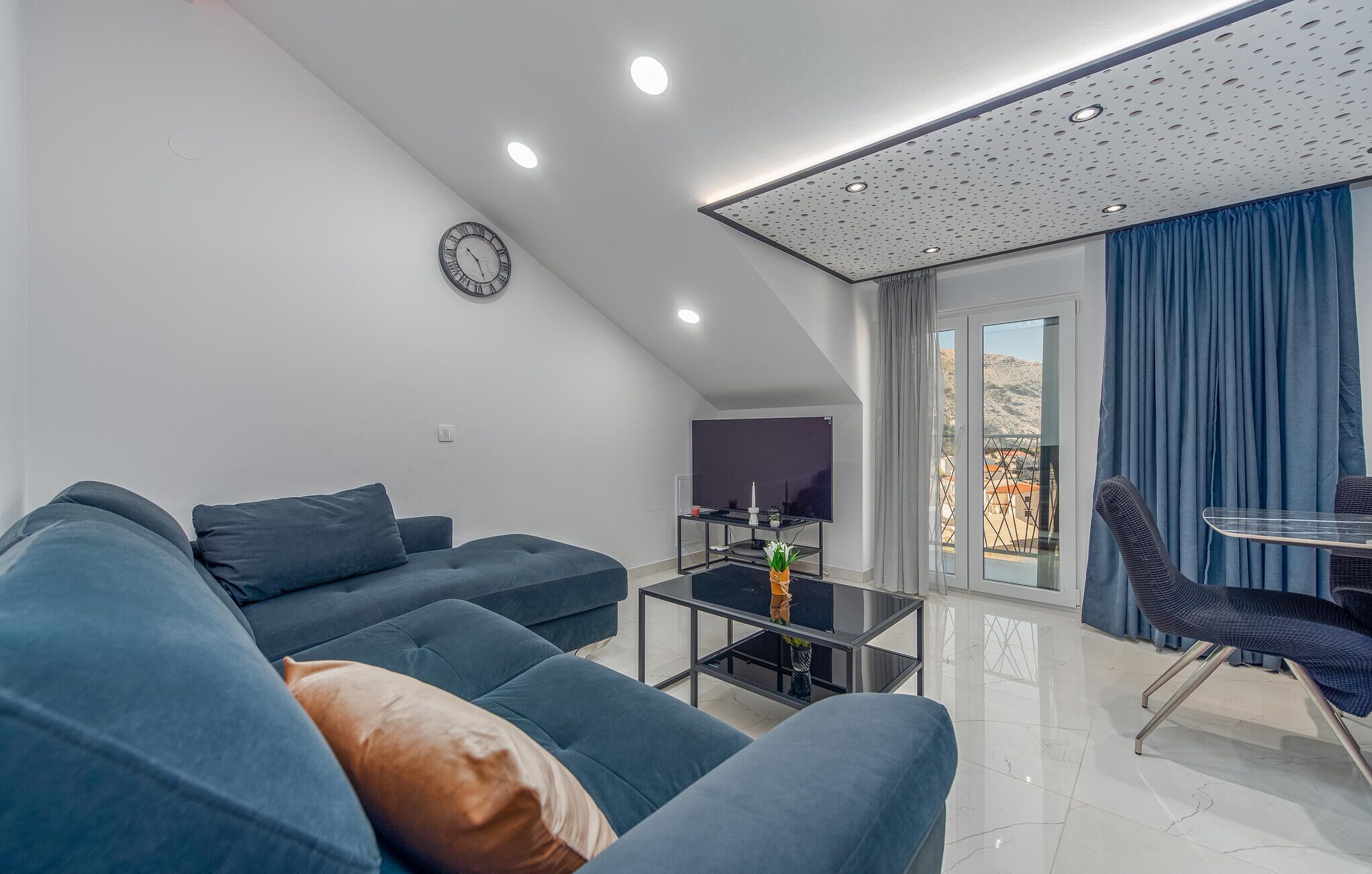 A modern living room with a sofa, a coffee table, and a TV. There is a clock on the wall and a balcony with curtains.
