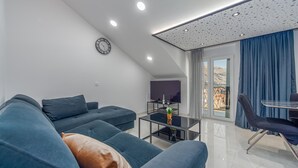 Un salon moderne avec un canapé, une table basse et un téléviseur. Il y a une horloge au mur et un balcon avec des rideaux.