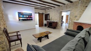Een ruime woonkamer met een stenen muur, een houten bank, een bankstel en een aan de muur bevestigde televisie.