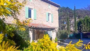 A stone building with green shutters, surrounded by lush greenery and a clear blue sky.