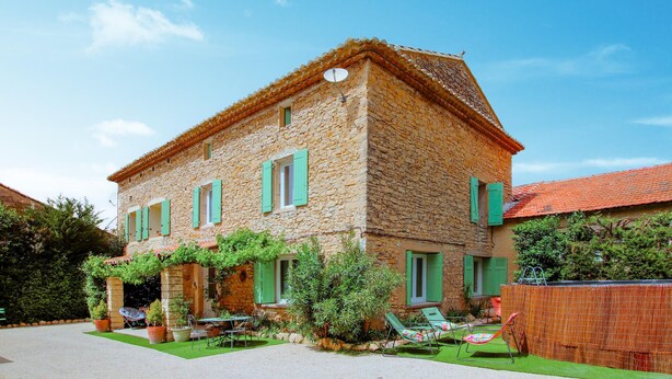 A stone building with a tiled roof, green shutters, and a small garden with outdoor seating.