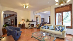 A spacious living room with a sofa, armchair, and glass coffee table. There is a fireplace, wooden cabinets, and a staircase leading to another level.