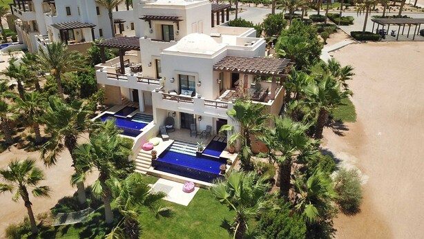 Een moderne meerlaags villa met zwembad, omgeven door palmbomen en een strand.