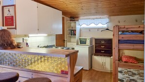 A compact kitchen with a wooden counter, a microwave, and a refrigerator. There is a bunk bed with a patterned mattress and a small shelf with books.