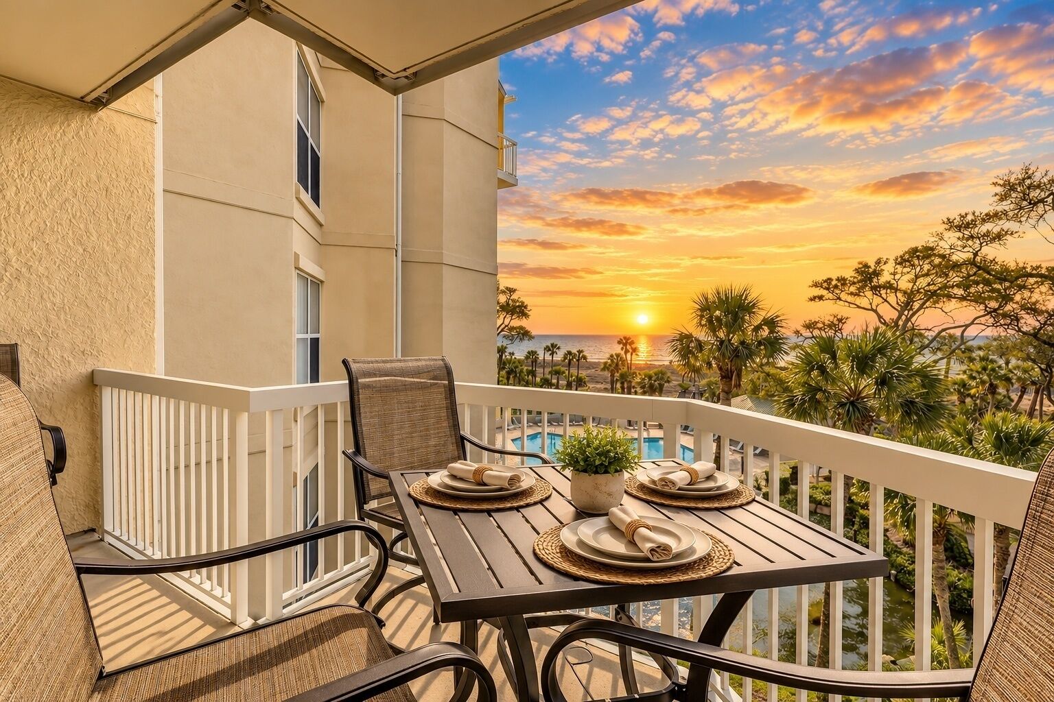 Un balcón con una mesa lista para dos personas, con vista a una piscina y palmeras.