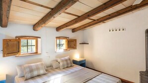 A bedroom with a wooden ceiling, two windows, a bed with checkered bedding, a nightstand, and a wall-mounted heater.