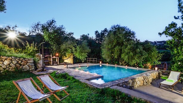 A rectangular swimming pool surrounded by stone walls, with two lounge chairs and a stone fire pit.