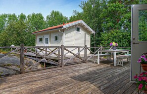 Une terrasse en bois mène à une petite cabane blanche au toit rouge, entourée d’une végétation luxuriante.