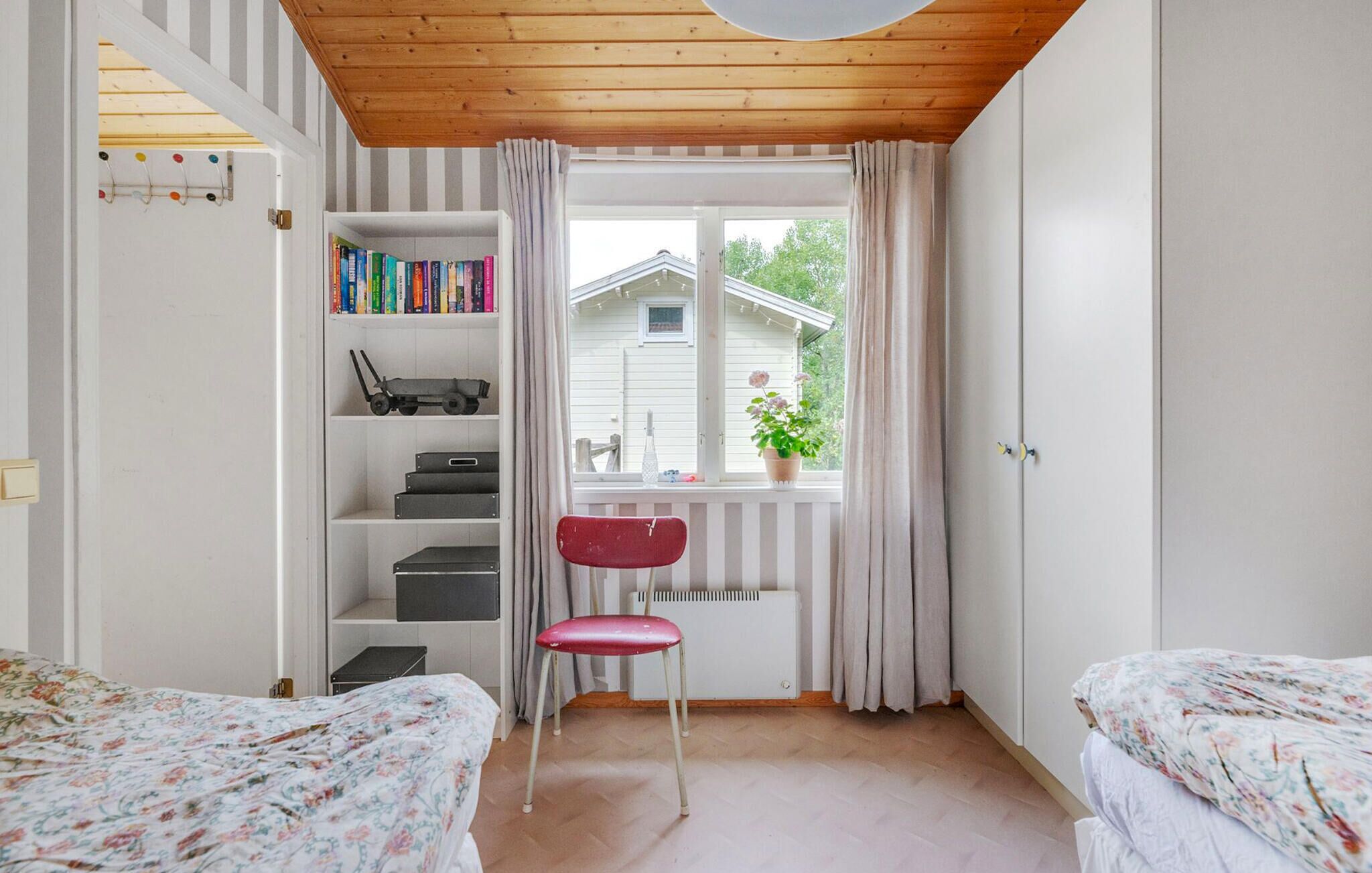 Un dormitorio con techo de madera, una ventana con cortinas, una estantería, un radiador y dos camas con ropa de cama estampada.