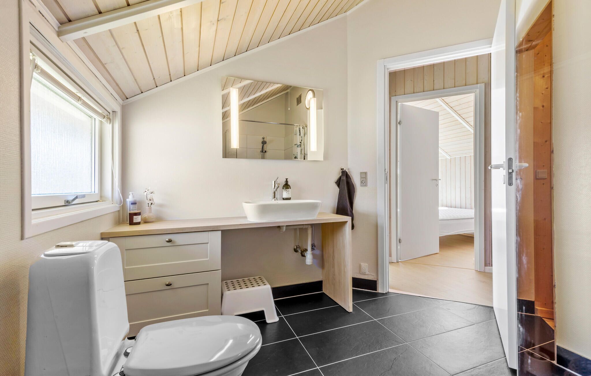Une salle de bain moderne avec des toilettes blanches, un lavabo et un miroir. Une fenêtre laisse entrer la lumière naturelle et une porte donne accès à une autre pièce.