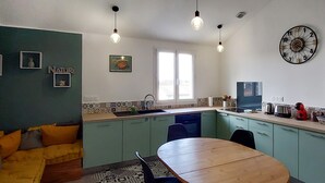 Une cuisine compacte avec un coin repas, comprenant un évier, une horloge et une fenêtre laissant entrer la lumière naturelle.