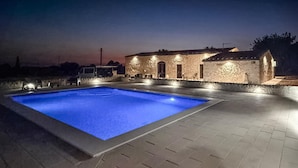 Una piscina con borde de baldosas, un edificio de piedra con varias ventanas y puertas, y una furgoneta estacionada.