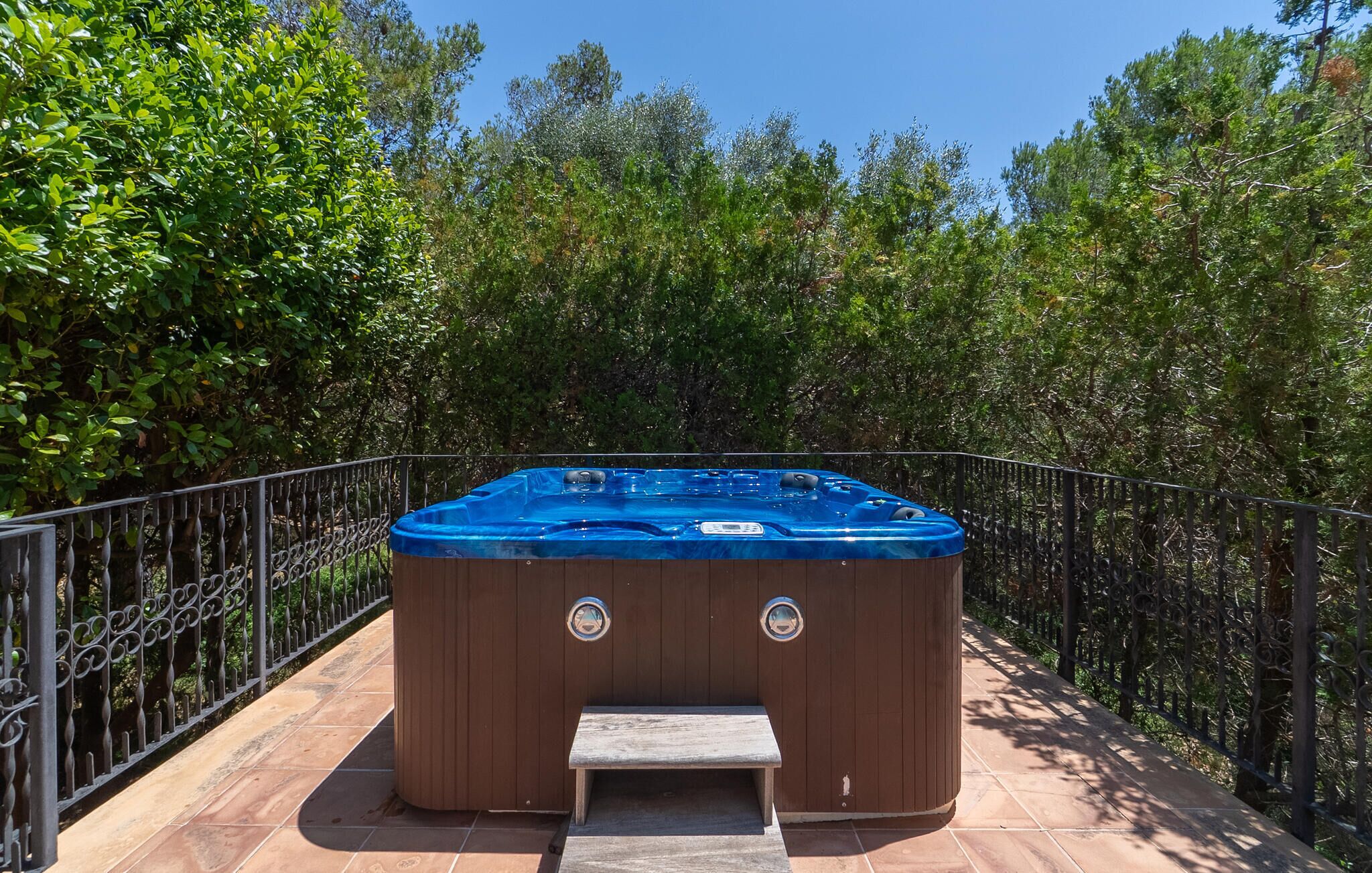 Een hottub met een houten buitenkant en een blauwe afdekking, omringd door een metalen hek en weelderige begroeiing.