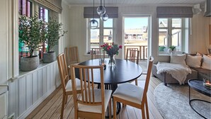 Uma sala de jantar com uma mesa redonda, cadeiras de madeira e um vaso de flores. Há plantas em vasos no parapeito da janela e vista para o exterior.