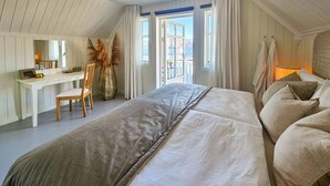 Quarto com cama grande, escrivaninha e cadeira. Há uma janela com cortinas e vista para o exterior.