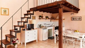 A compact kitchen with a dining area, a staircase with a TV on the landing, and a small refrigerator.